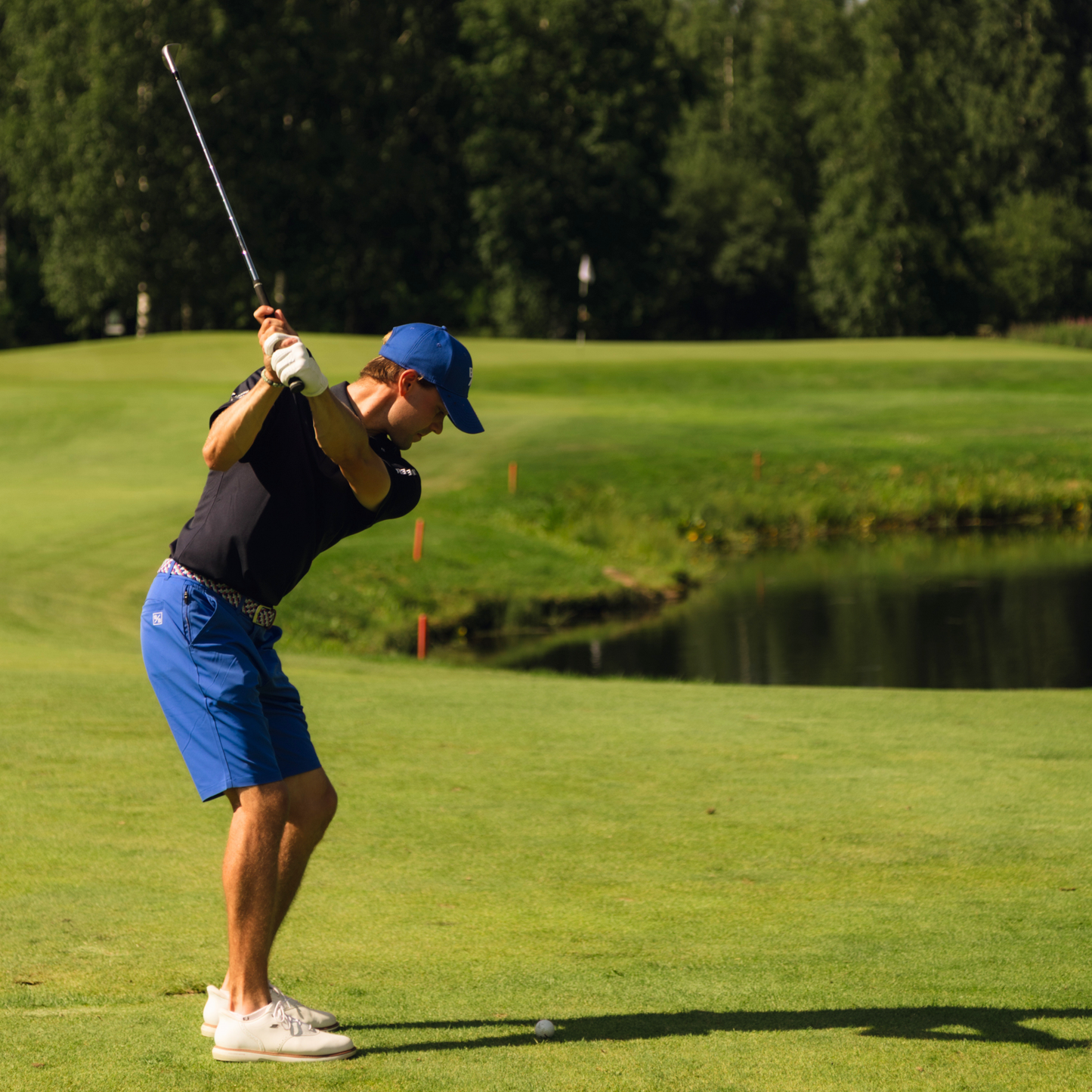 A golfer in bright blue golf shorts, and a blue cap is captured mid-swing on a golf course 