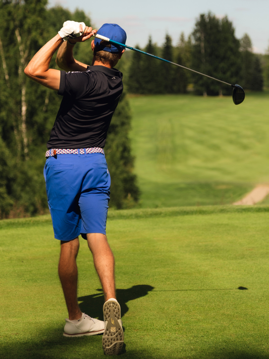 A man on a golf course mid-swing, wearing a black polo shirt, bright blue golf shorts, a blue cap, and white golf shoes.